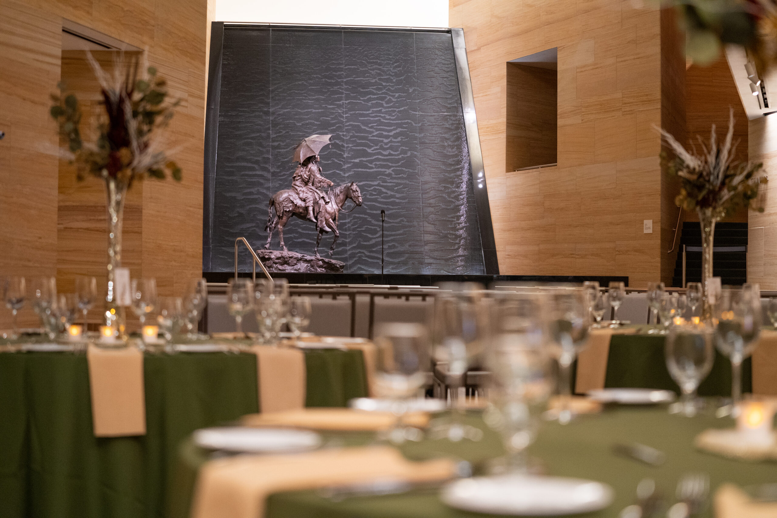 Tables set for an event with green tablecloths and glassware in the foreground highlight the elegance of our Event Spaces; a statue of a person on horseback stands before a water feature in the background.
