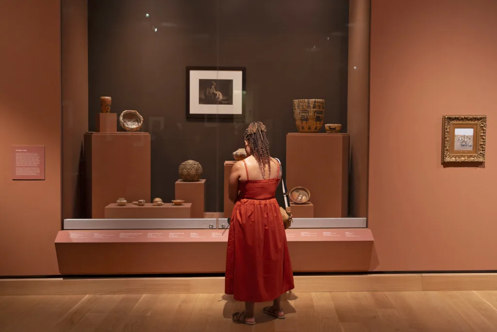 A woman in a red dress stands in front of a glass museum display case containing pottery and baskets in the Early West Gallery, with framed artwork on the brown walls.