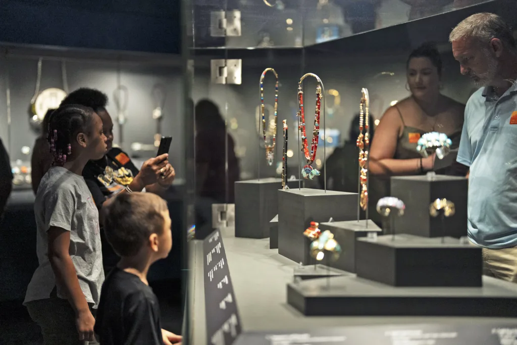 Several people, including children and adults, view and photograph jewelry pieces displayed in glass cases at the museum's Jewel Box exhibit.