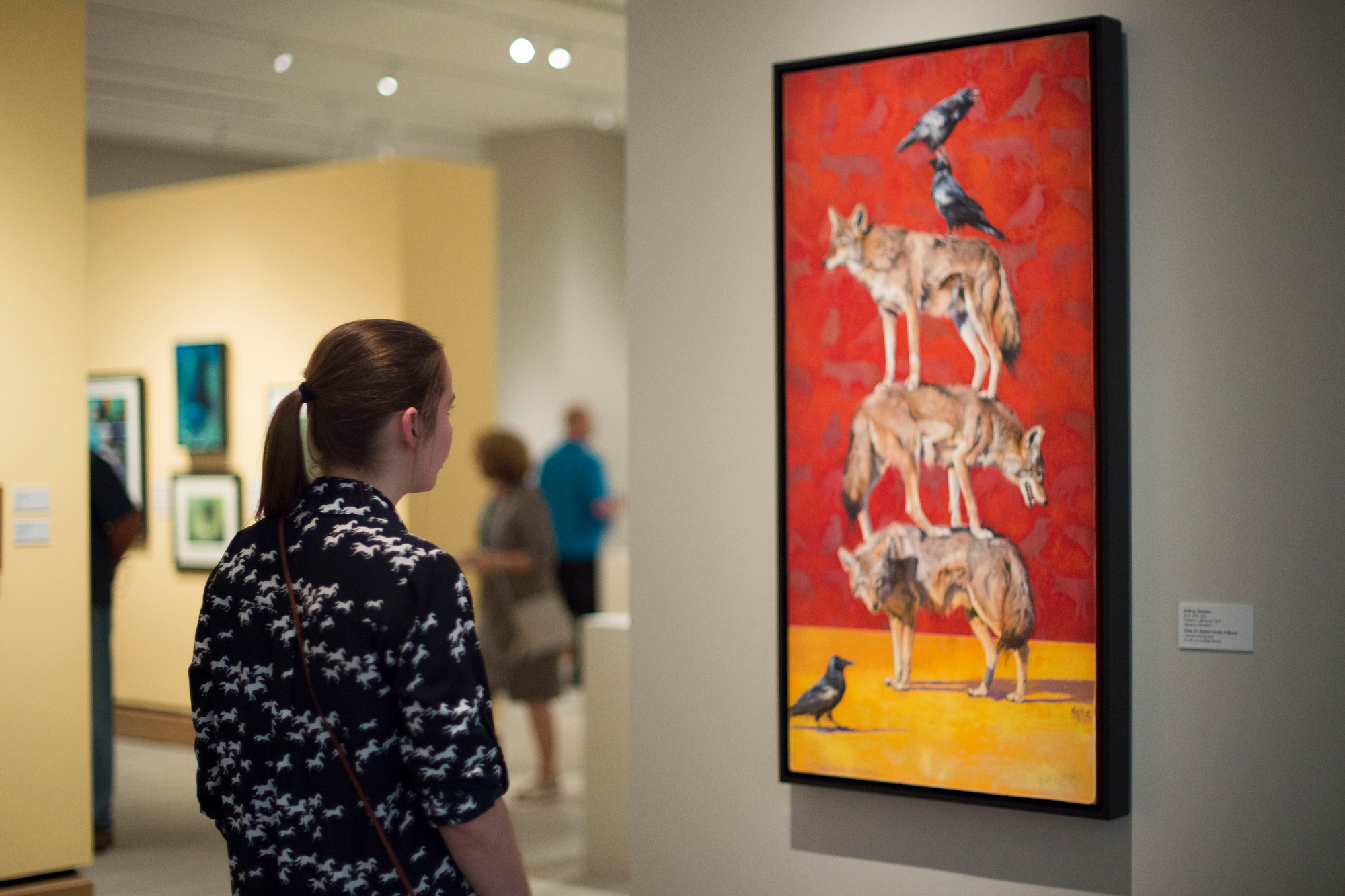 A woman views a painting of three stacked coyotes, butterflies, and several black birds in an art gallery.