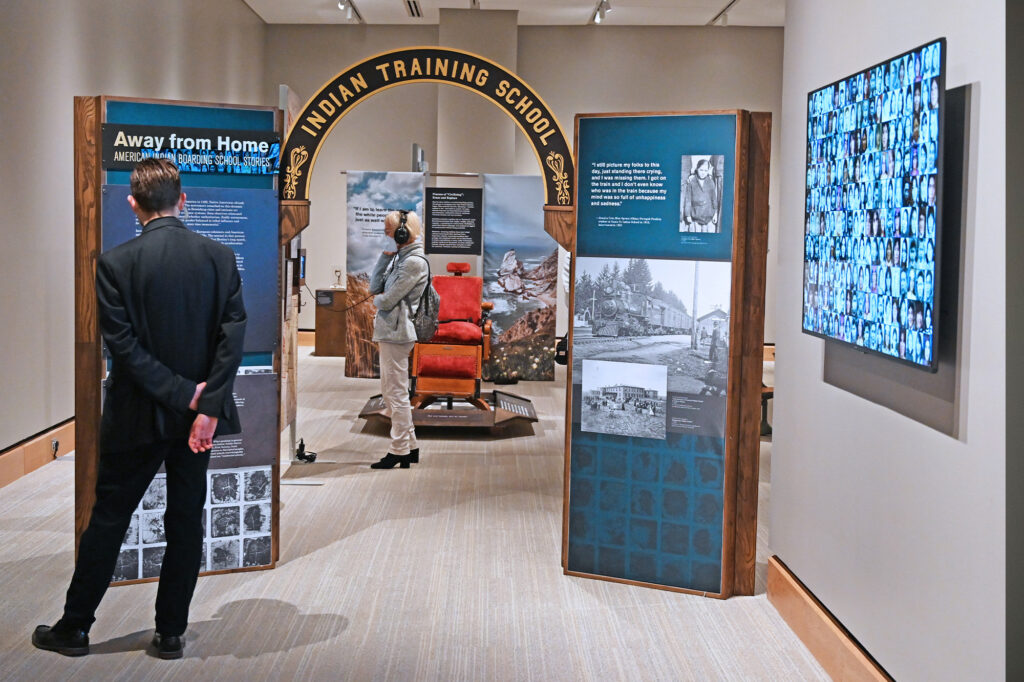 A person views an exhibit titled "Indian Training School" with informational panels, photos, and a display of historical images on a screen, alongside references to local endangered birds.