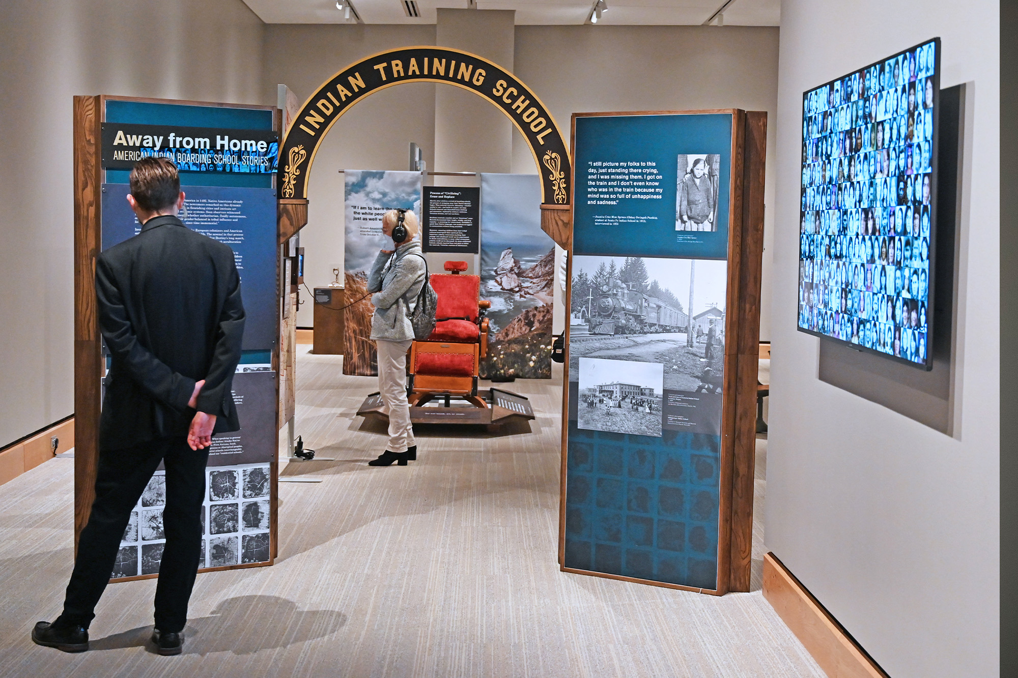 A person views an exhibit titled "Indian Training School" with informational panels, photos, and a display of historical images on a screen, alongside references to local endangered birds.
