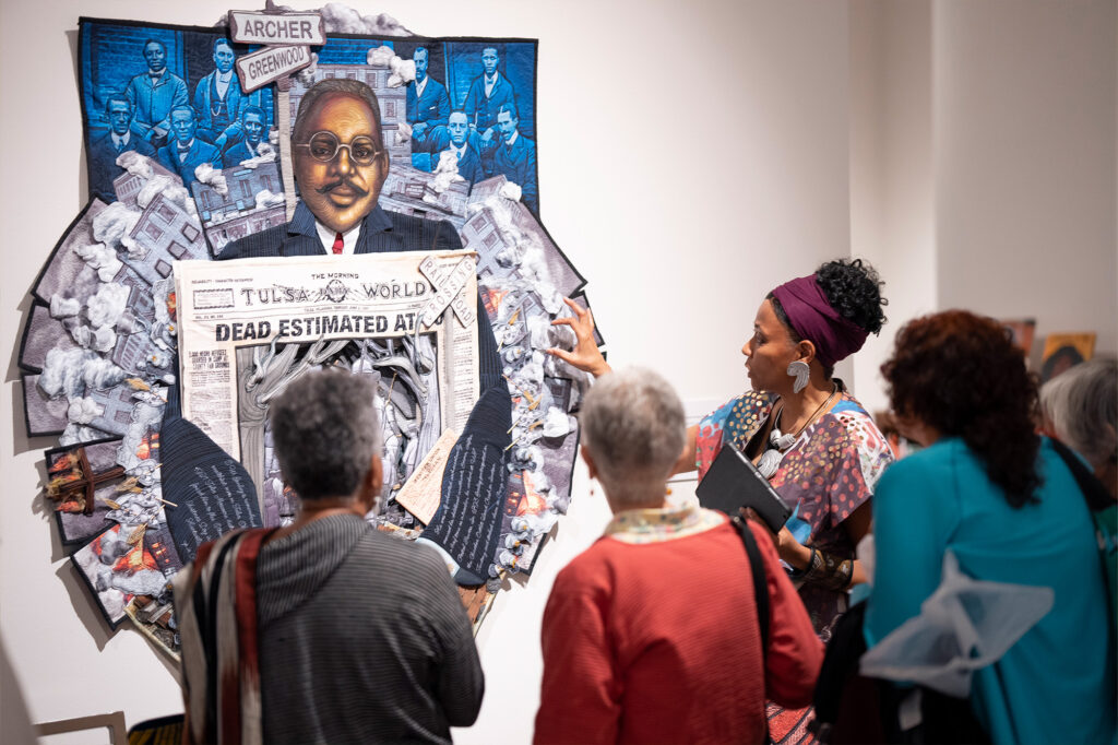 A woman explains an artwork depicting a historical figure surrounded by endangered birds and newspaper headlines to a group of people at an art exhibit.