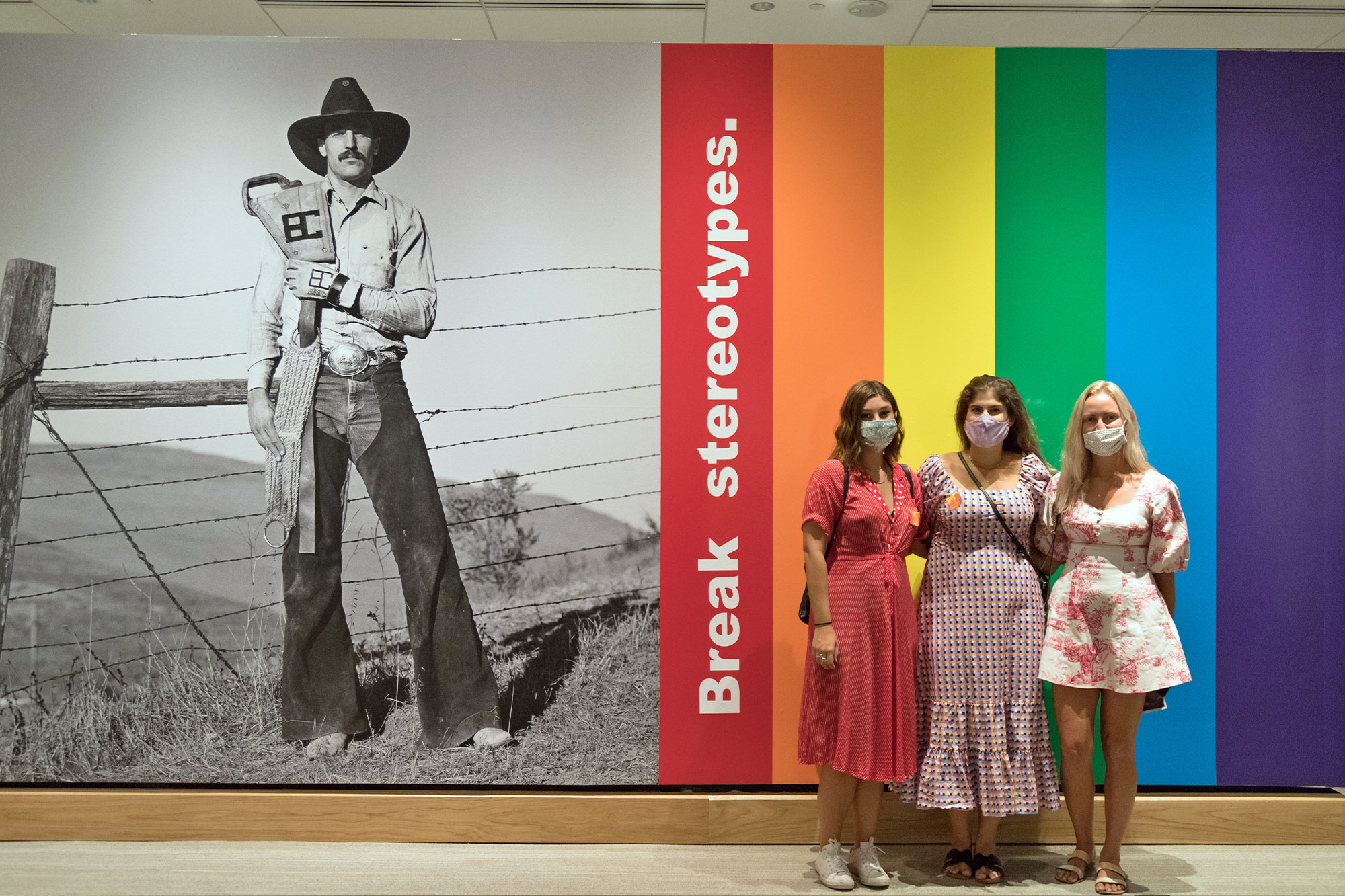 Three women in dresses and masks stand in front of a rainbow-colored wall, alongside the text "Break stereotypes" and a large black-and-white photo of a cowboy, with butterflies accenting the vibrant scene.