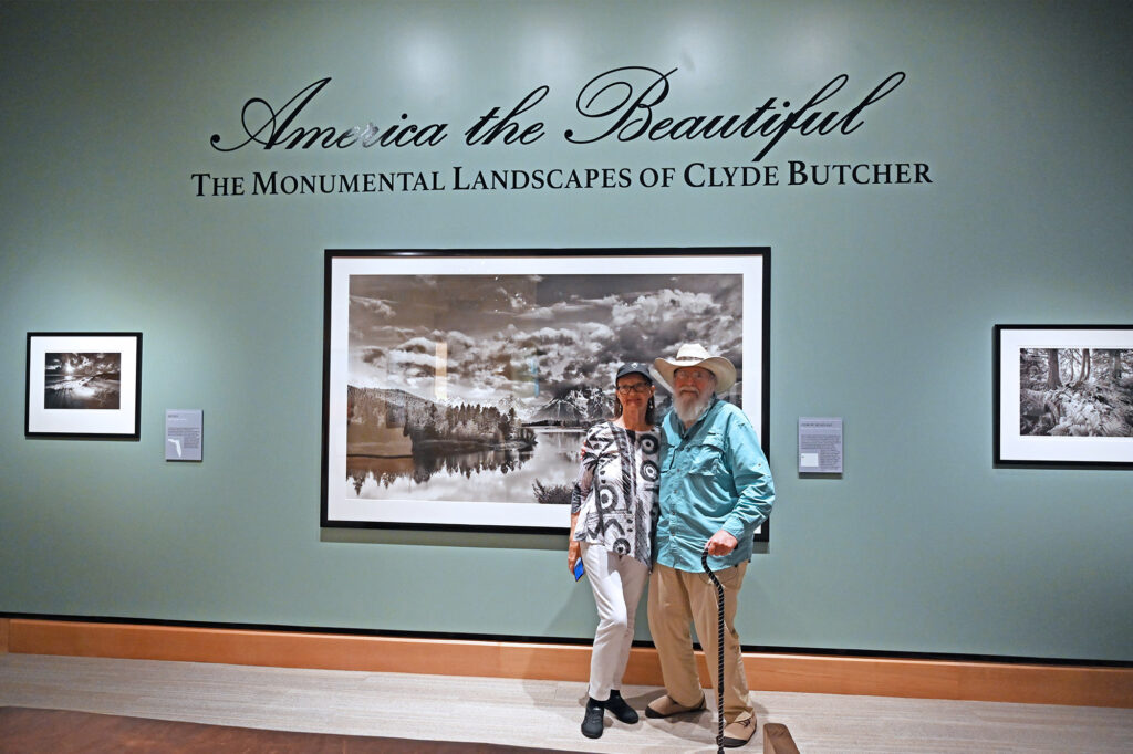 Two people stand in front of framed landscape photos, some featuring endangered birds, in an art exhibit titled "America the Beautiful: The Monumental Landscapes of Clyde Butcher.