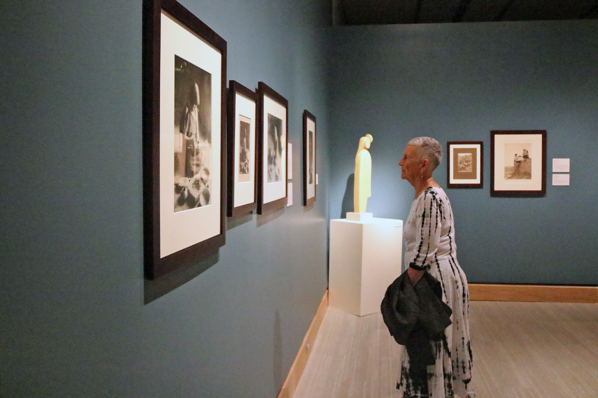 An older woman in a black and white dress admires framed photographs of endangered birds on display in an art gallery with blue walls.