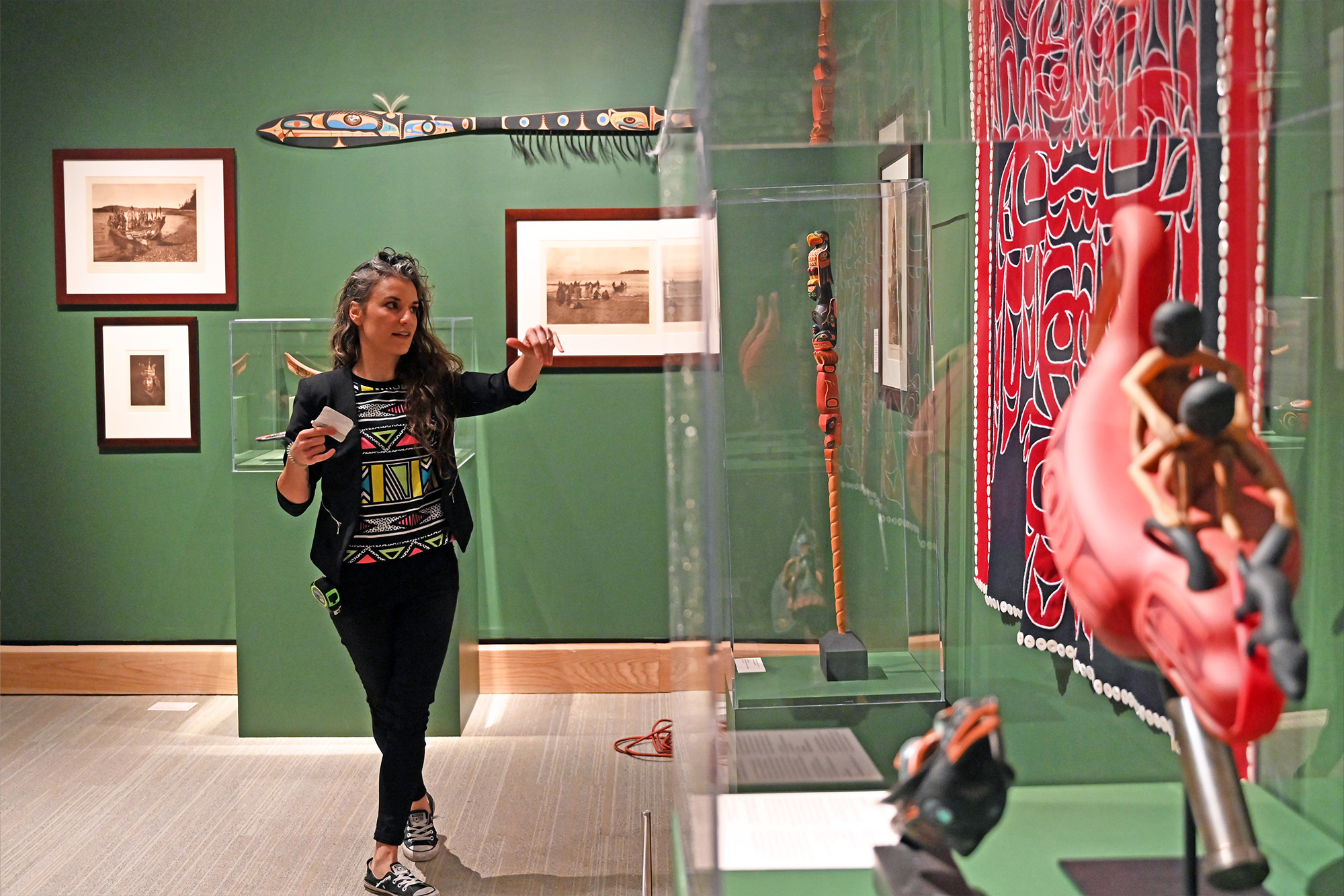 A woman walks through an art gallery, viewing Indigenous artworks and artifacts—some inspired by endangered birds—displayed in glass cases and hung on green walls.