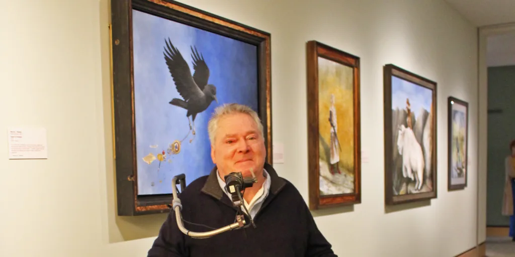 A man stands at a microphone in front of a wall displaying framed paintings from Marcus C. Thomas’s "Flight of the Mind" collection in an art gallery.