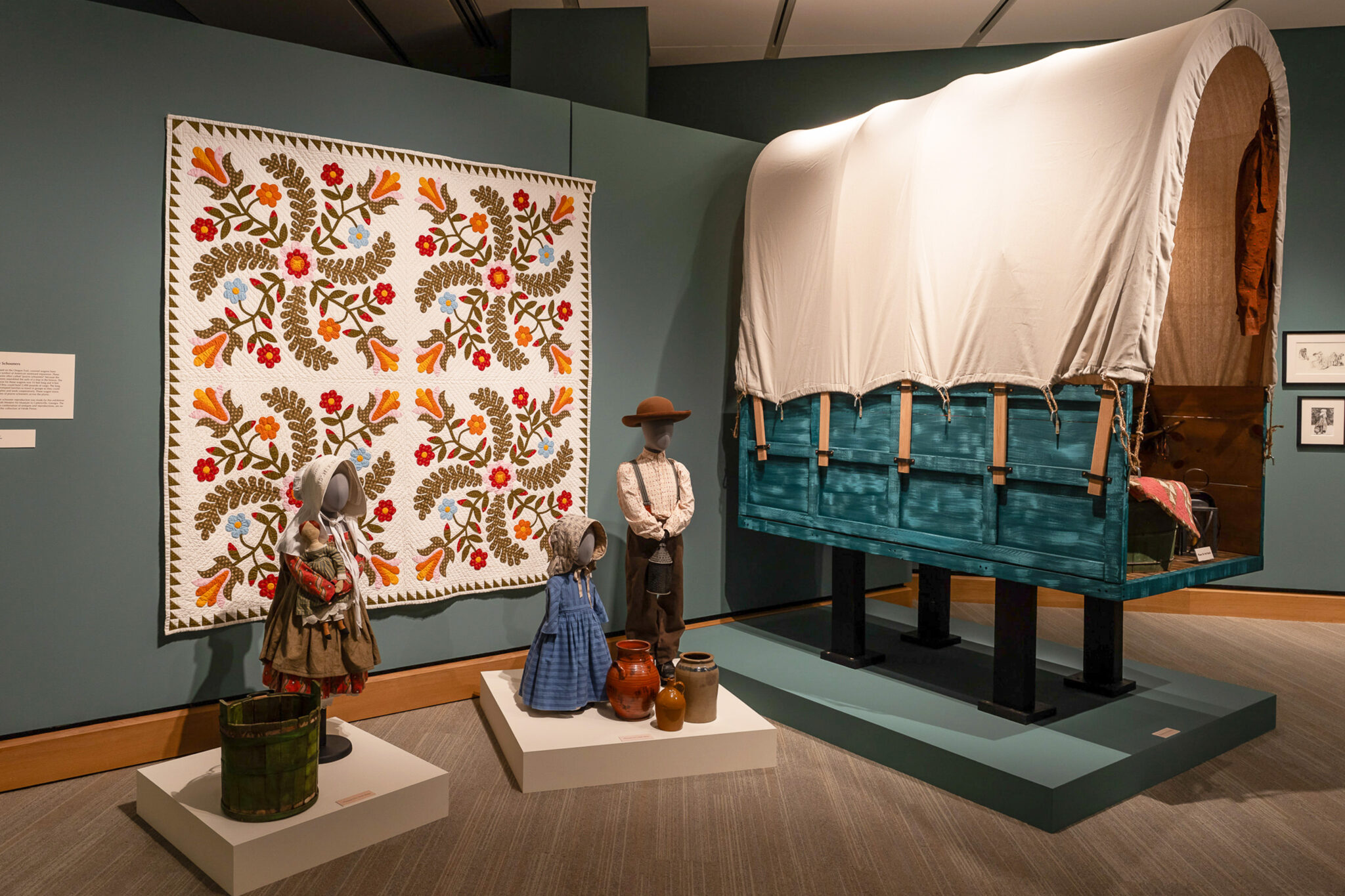A quilt hangs on the wall beside a covered wagon display, with three mannequins dressed in pioneer clothing and several clay pots on the floor, recalling a time when endangered birds still filled the skies above.