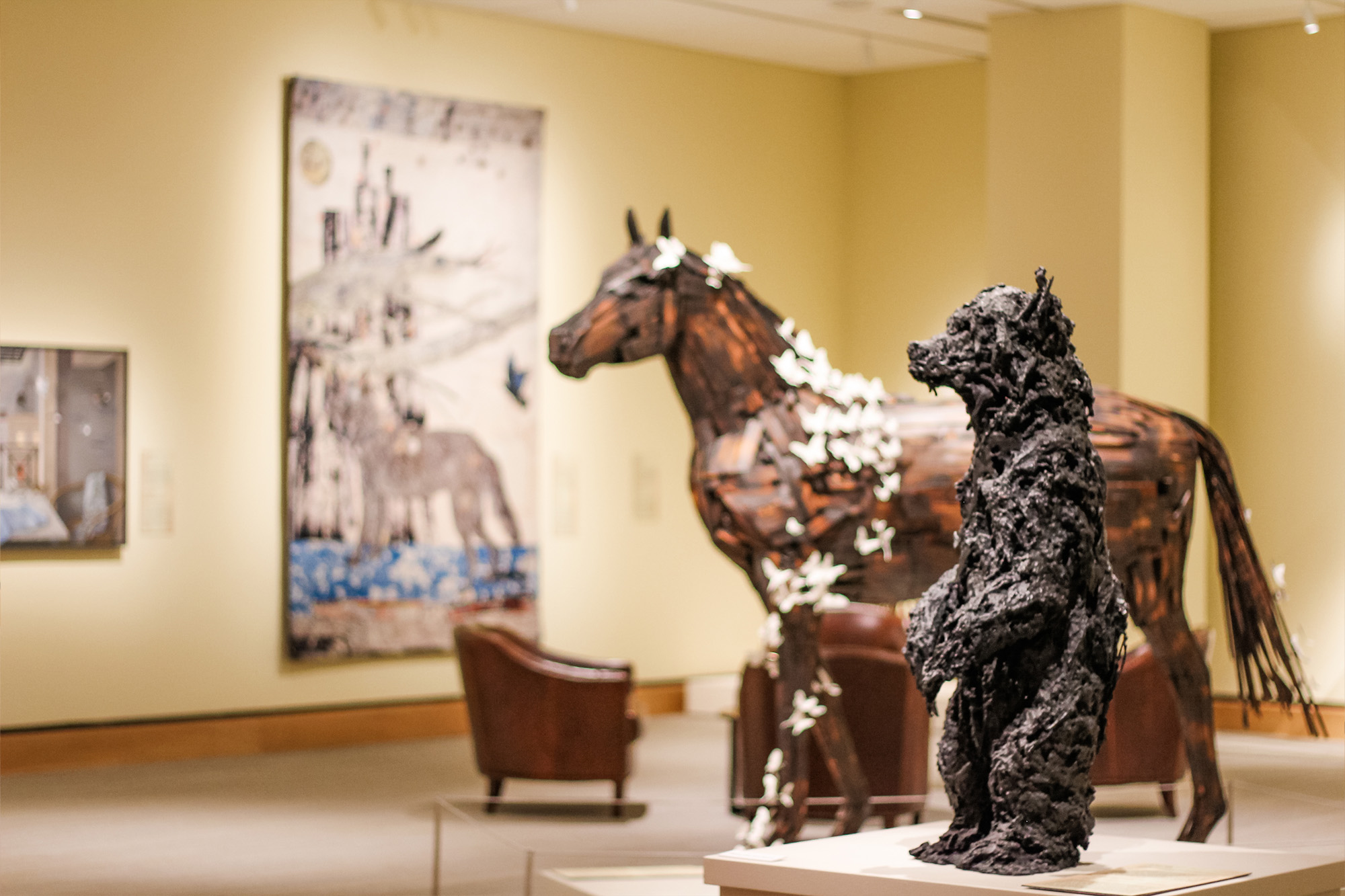 A museum gallery displays a bear sculpture in the foreground, a horse sculpture with endangered butterfly accents, and various artworks on the walls.