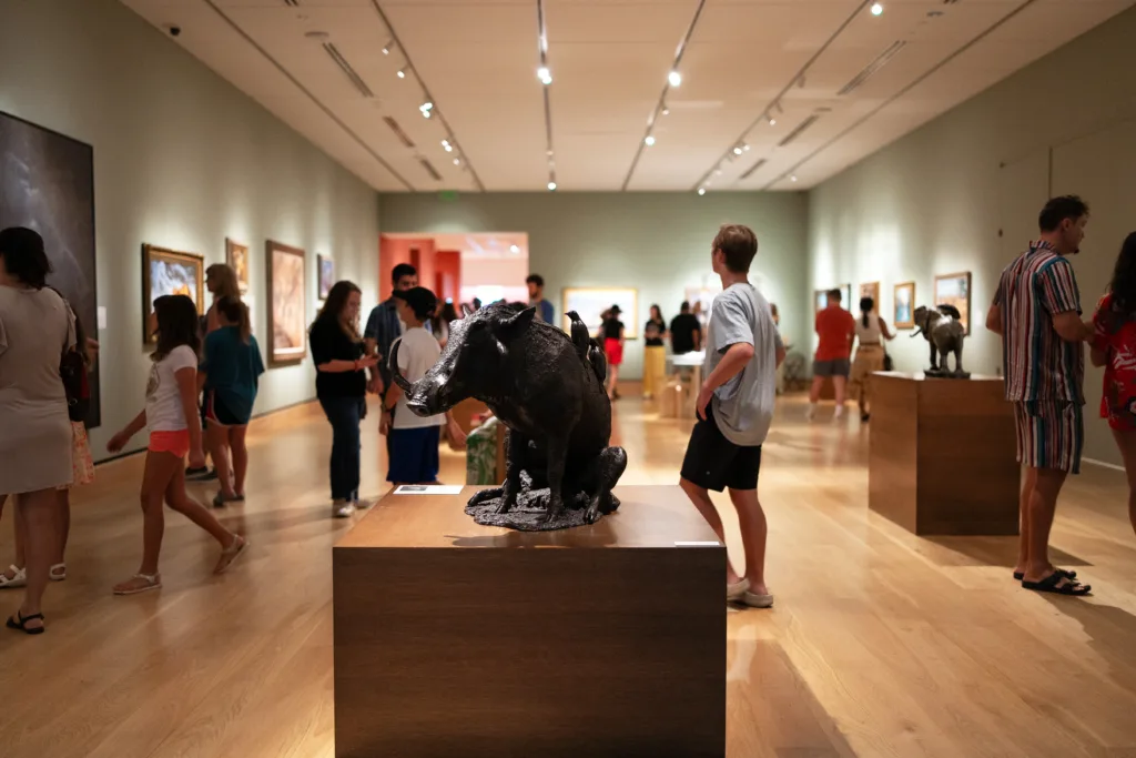 People walk around and view artwork and wildlife sculptures in the well-lit Wildlife Gallery, featuring wood floors and green walls, curated by the Earth & Humanity Foundation.