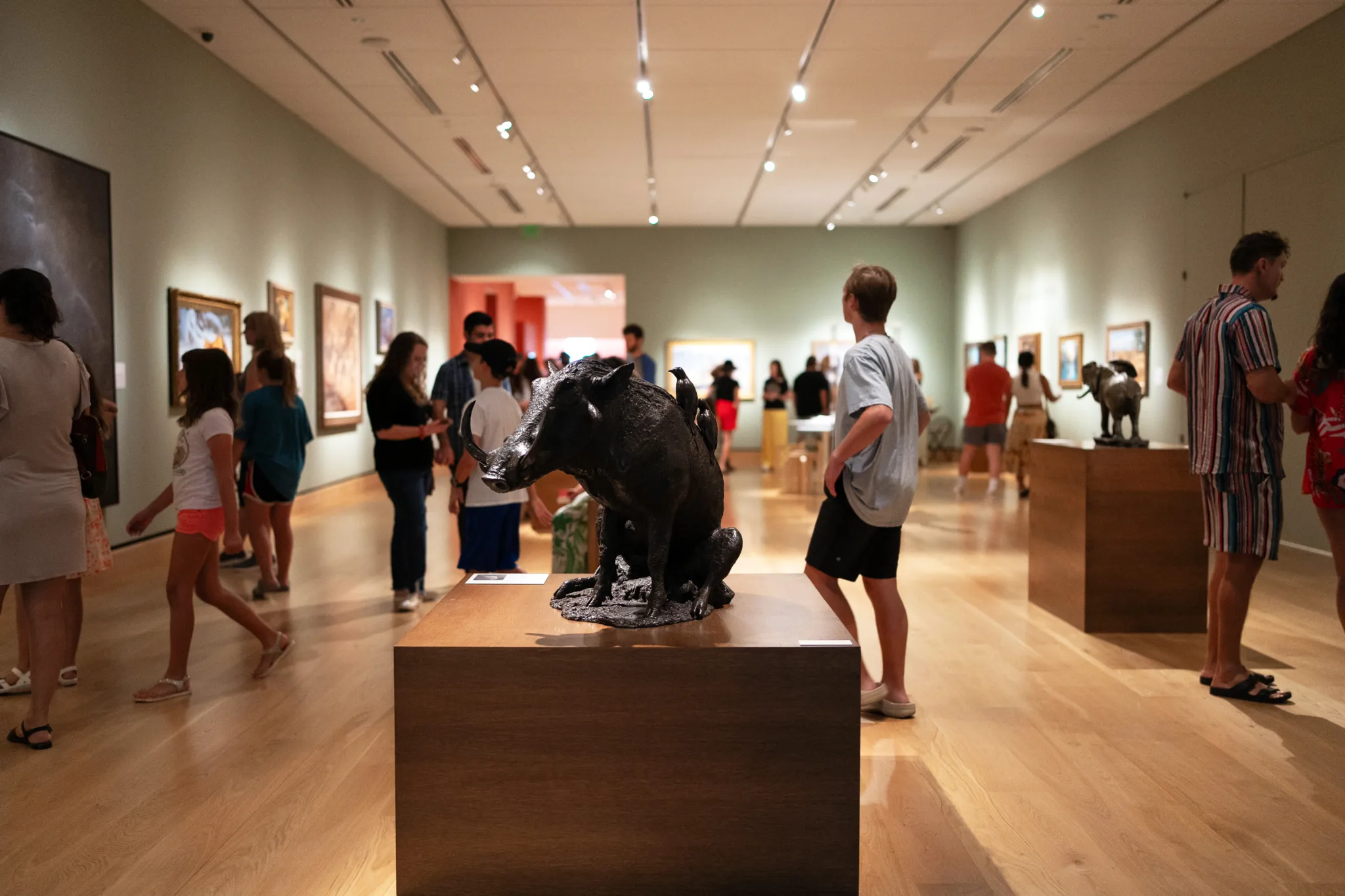 People walk around and view artwork and wildlife sculptures in the well-lit Wildlife Gallery, featuring wood floors and green walls, curated by the Earth & Humanity Foundation.