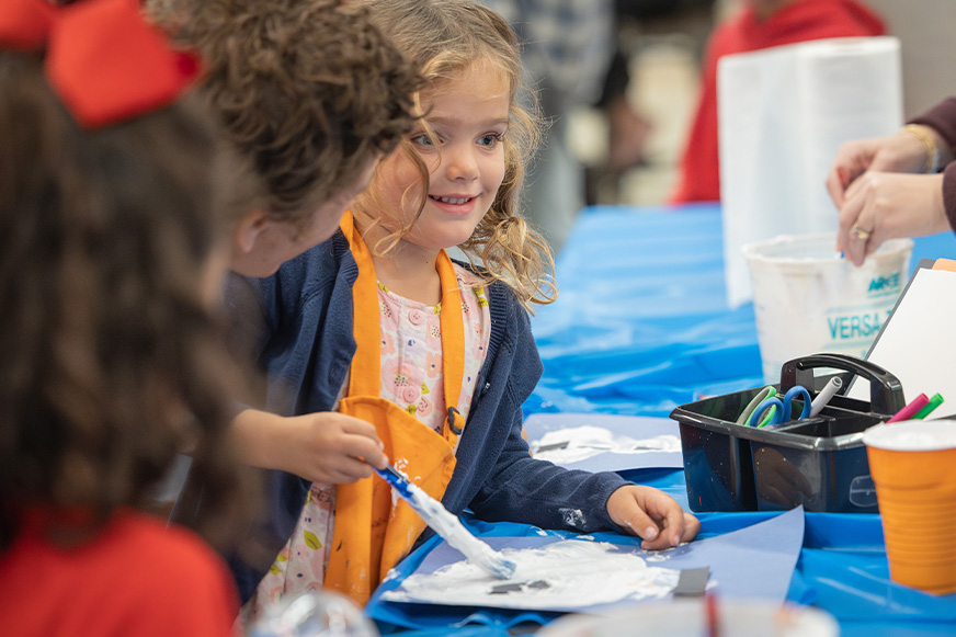 A young girl smiles while painting at a table covered with blue paper during Family Artmaking, surrounded by art supplies and other children.