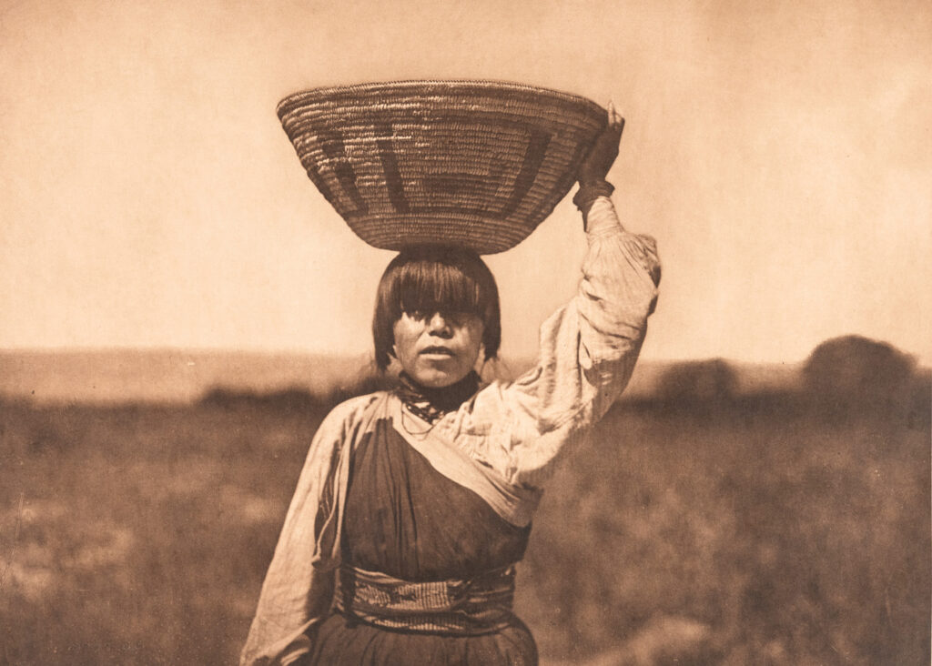 A young person stands outdoors, balancing a large woven basket on their head with one hand, in a scene reminiscent of photographer Edward S. Curtis, set against a blurred natural background.