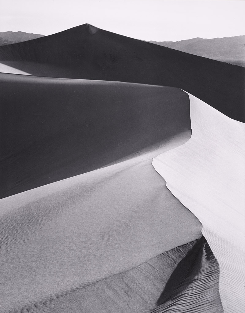 Black and white photography of sand dunes with sharp ridges and shadows, evoking the masterworks of Ansel Adams through striking contrasts in a dramatic desert landscape.