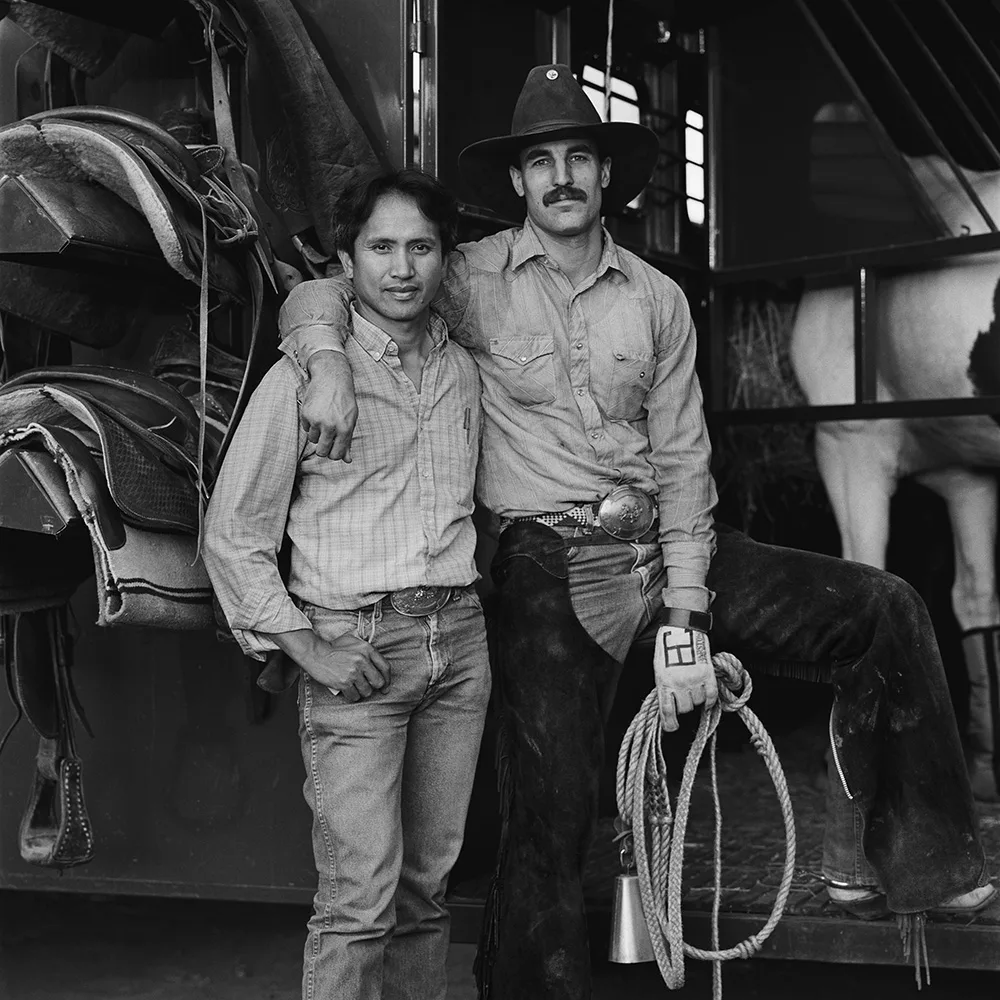 Two men stand side by side in front of a horse trailer; one, dressed as a cowboy holding a rope, appears ready for the Gay Rodeo. Saddles are visible to the left in this striking Blake Little photograph.