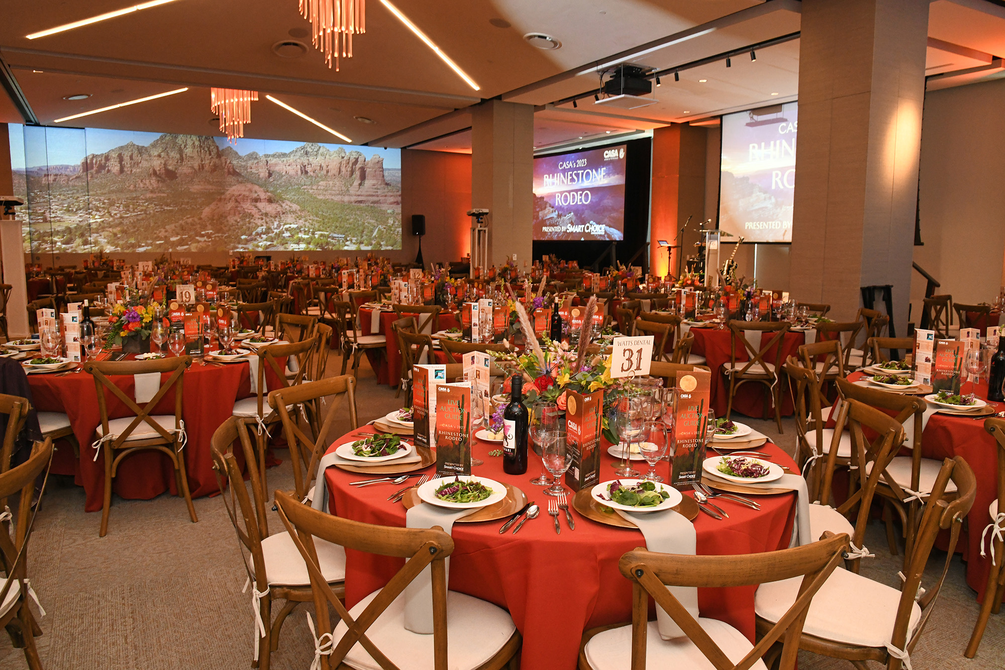 A banquet hall with round tables set for dinner, featuring red tablecloths, salads on plates, floral centerpieces, and large screens displaying event graphics—a perfect example of elegant Event Spaces.