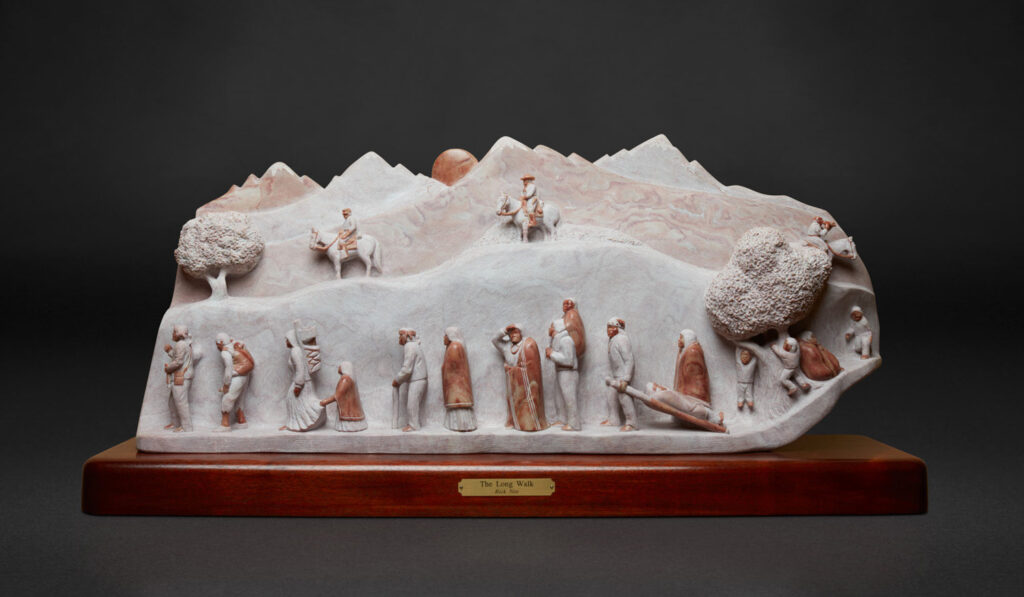 Stone sculpture in the style of Western Art, depicting a group of people and animals traveling along a winding path in a mountainous landscape, mounted on a wooden base labeled "The Long Walk.
