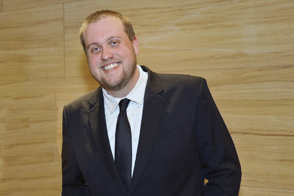 A man wearing a black suit, white shirt, and black tie stands smiling in front of a light wood-paneled wall.