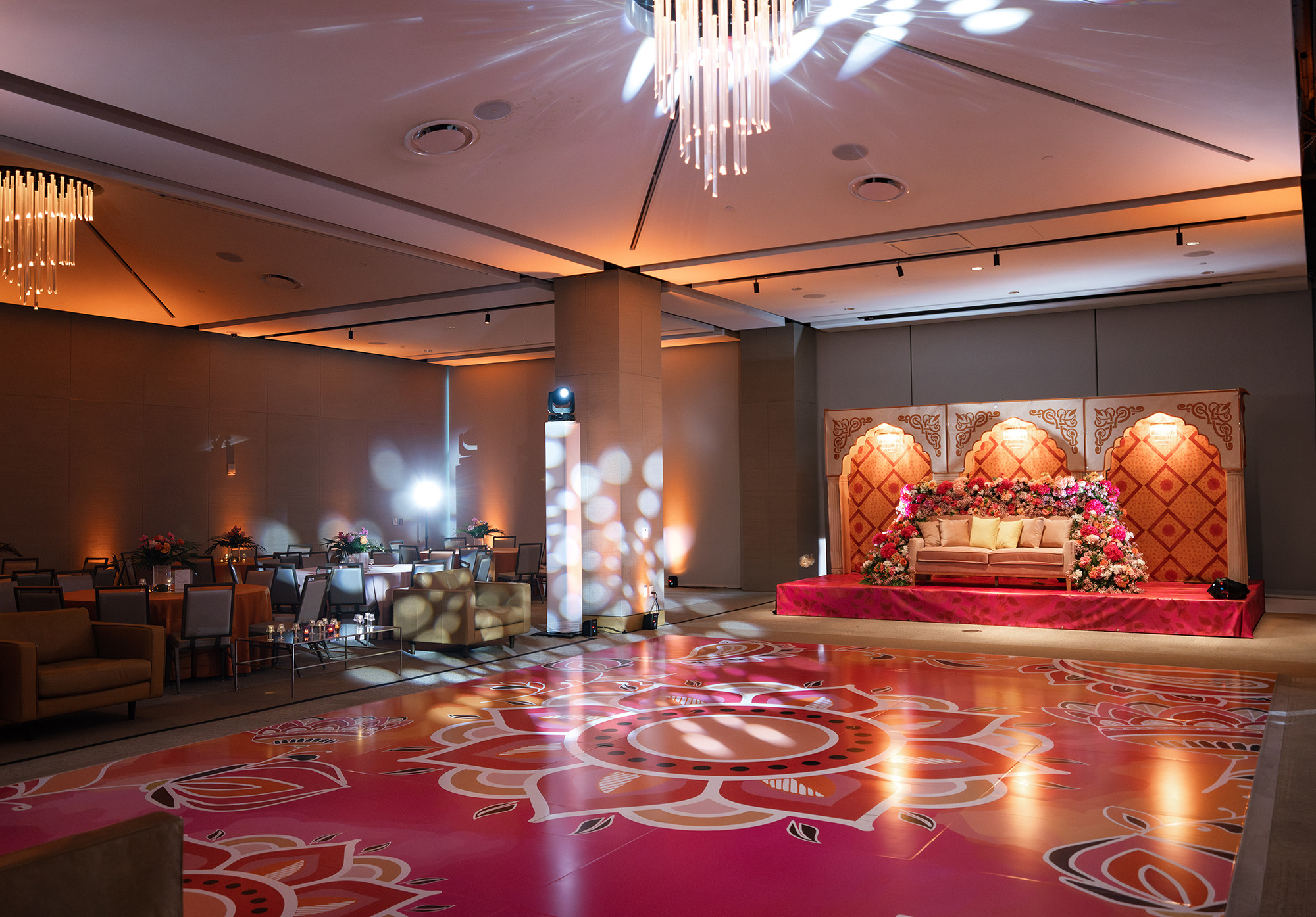 A decorated banquet hall event space with a patterned pink dance floor, a stage adorned with flowers and a sofa, round dining tables, and modern chandeliers overhead.