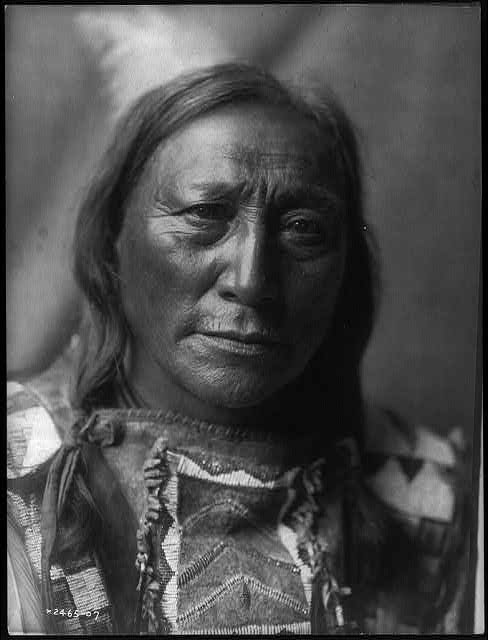 Black and white portrait of a Native American man with long hair, wearing a beaded shirt and looking directly at the camera, evoking a sense of mystery archive.
