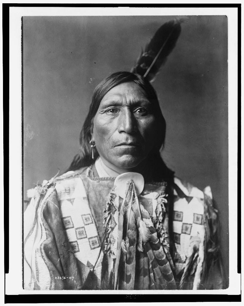 Black-and-white portrait of a Native American man in a beaded shirt and traditional clothing, with a feather in his hair, facing forward against a plain background, evoking an air of mystery.