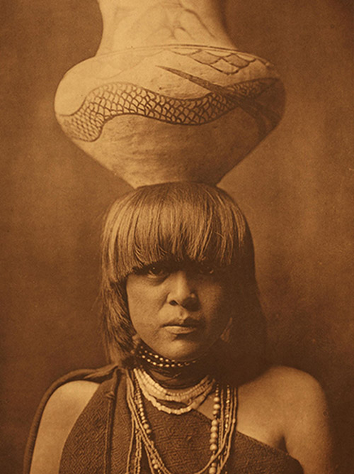 A woman with straight hair and layered necklaces balances a large, patterned ceramic vessel on her head, capturing a moment reminiscent of Indigenous Art photography in the style of Edward S. Curtis.