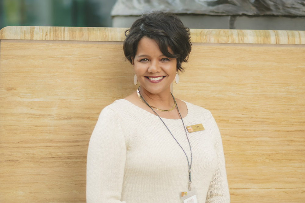 A woman with short dark hair wearing a cream sweater and name badge smiles while standing in front of a tan stone wall.