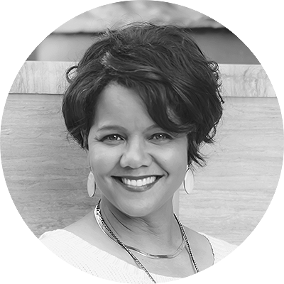 Smiling woman with short, wavy dark hair, wearing earrings and a necklace, posed in front of a light-colored background often found in elegant event spaces.