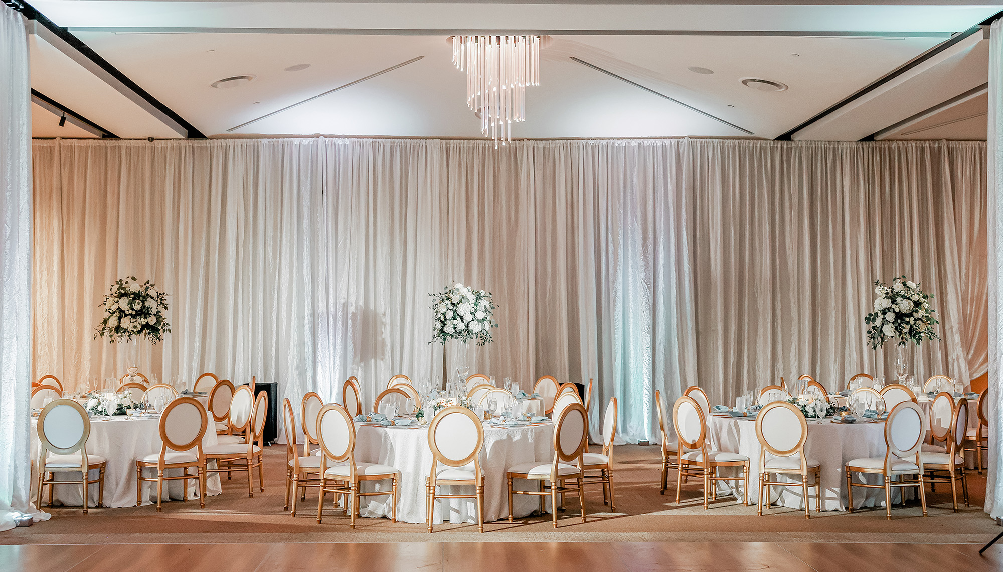A formal event hall, perfect for Event Spaces, features round tables set for dining, white chairs with gold trim, elegant floral centerpieces, and beige drapes.