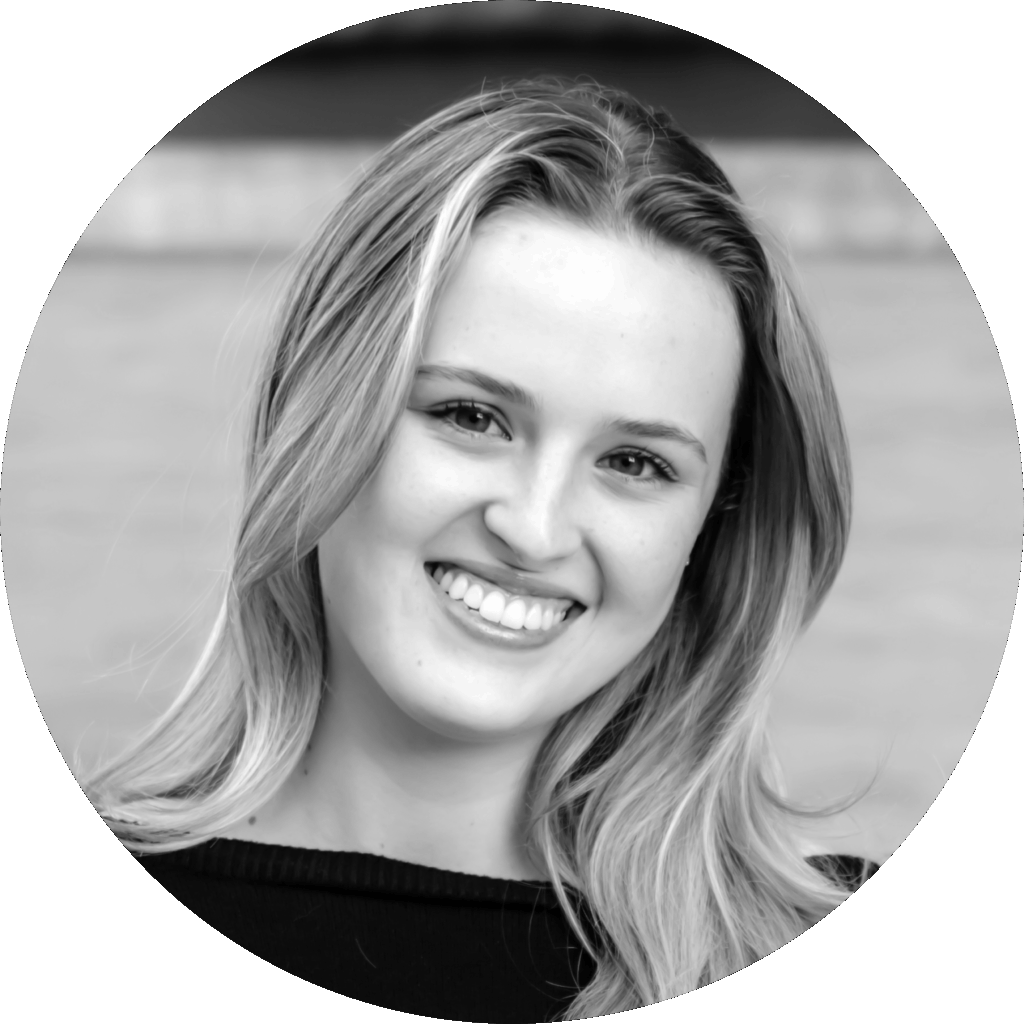Black and white headshot of a young woman with long hair, smiling at the camera against a blurred background often seen in vibrant event spaces.