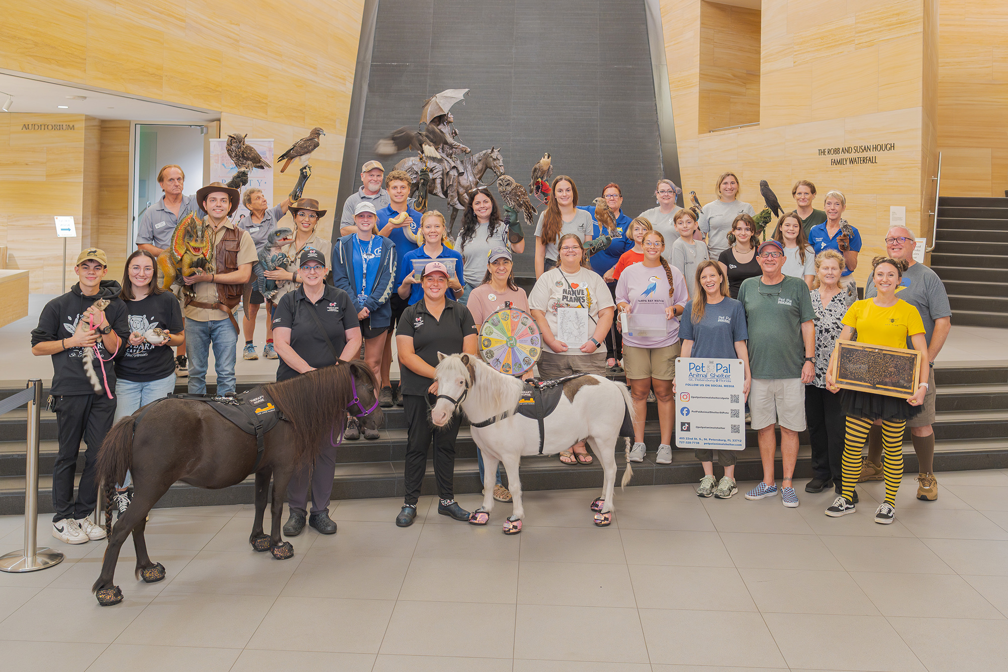 A large group of people, joined by their partners, pose indoors with two miniature horses, birds, and various props in front of a statue and stairs.