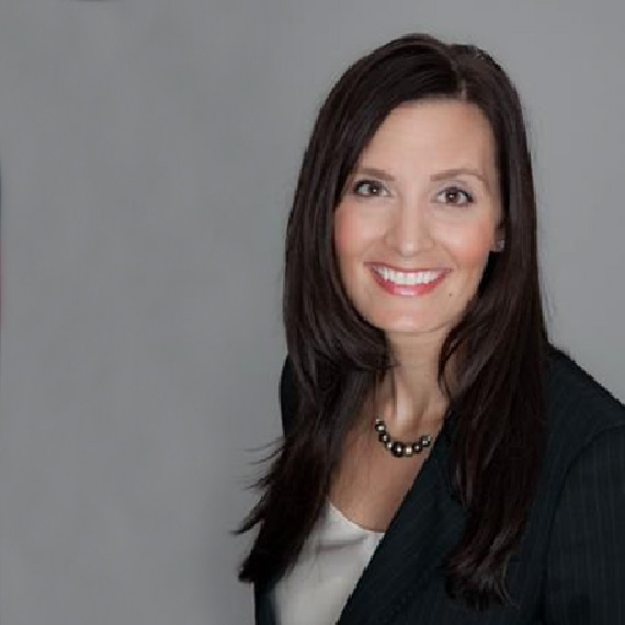 A woman with long brown hair, wearing a black blazer, white top, and a beaded necklace, smiles at the camera against a plain gray background.