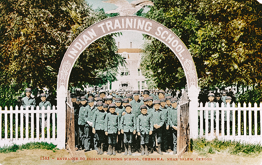 A group of boys in uniform stand under an arch labeled "Indian Training School" at the entrance to Chemawa, an American Indian Boarding School near Salem, Oregon; white fence and building in background.