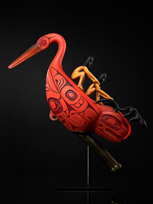 A red sculpted bird with Indigenous-style patterns, inspired by Indigenous Art, carries three human figures on its back, displayed against a black background.