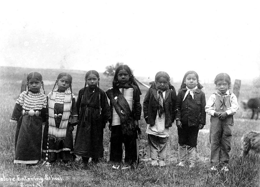 A group of seven Sioux children, some in traditional American Indian clothing and others in Western attire, stand in a row outdoors on grass, reflecting the stories of cultural change linked to the boarding school era.
