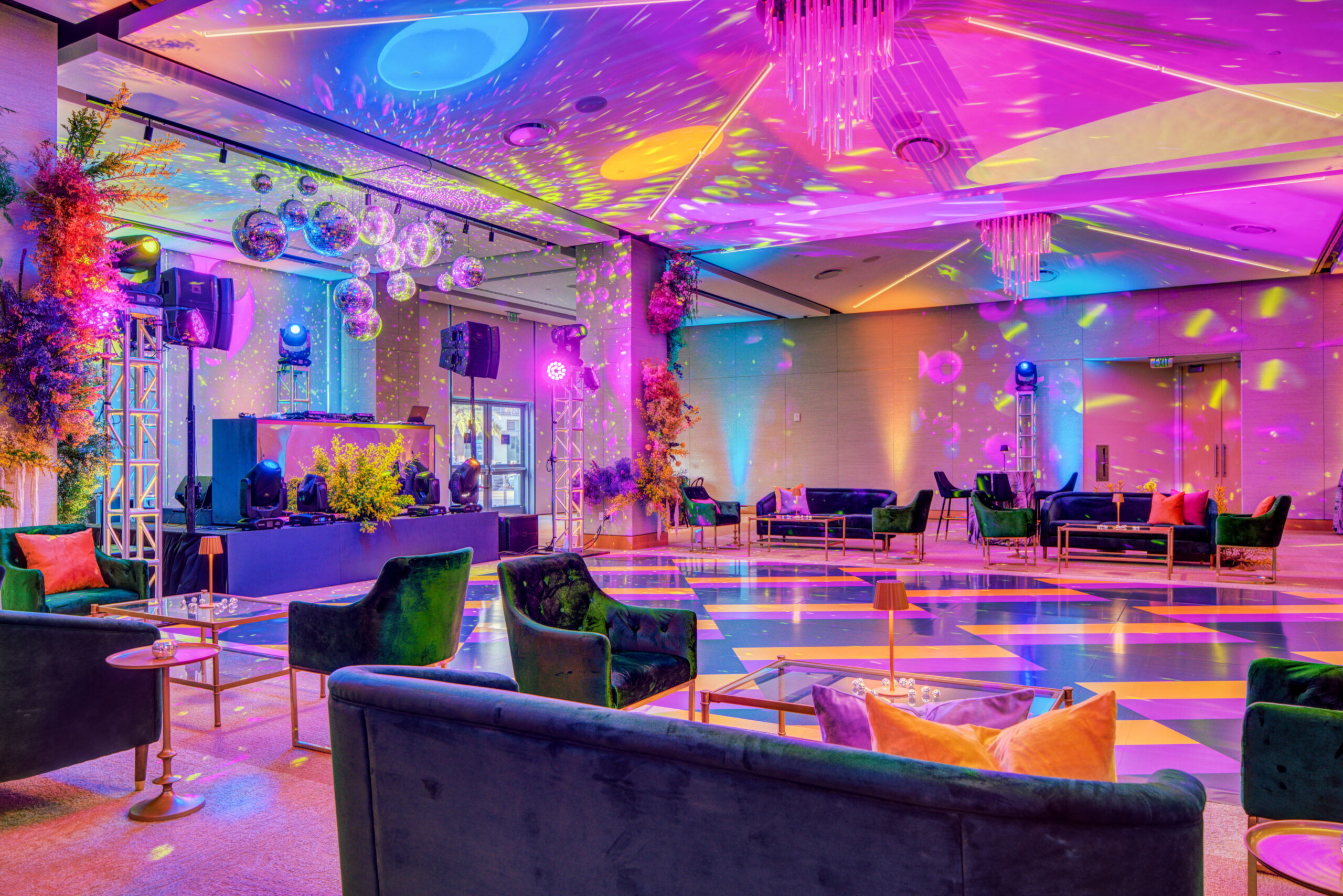 A modern event space with velvet seating, a checkered dance floor, and colorful lighting effects illuminating the room’s ceiling and walls. Disco balls and a DJ booth complete this standout choice among Event Spaces.