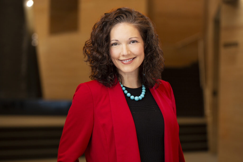 Woman with curly brown hair wearing a red blazer, black top, and turquoise beaded necklace, smiling while standing indoors with warm lighting and blurred background.