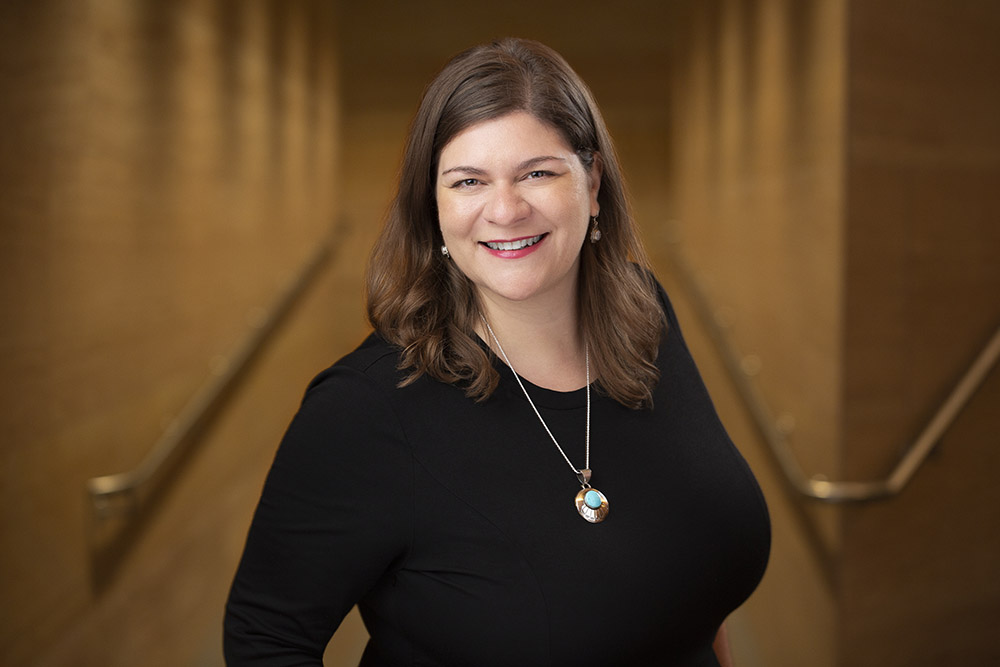 A woman with brown hair wearing a black top and a pendant necklace smiles at the camera, standing in a warmly lit hallway with gold-colored walls.