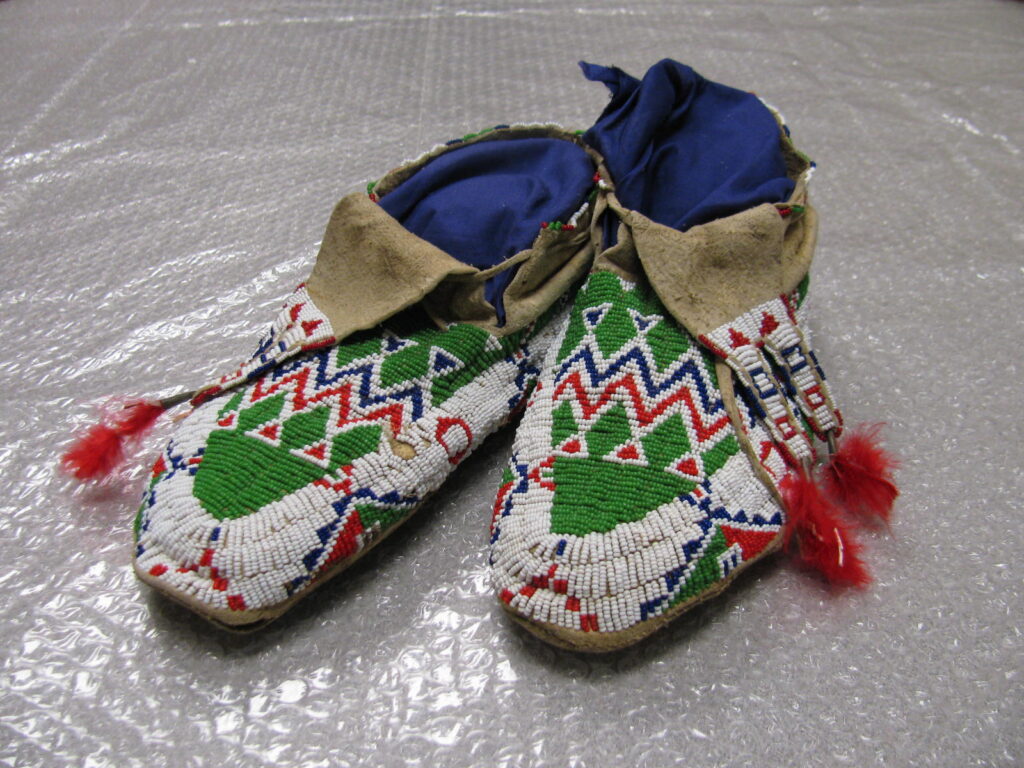 A pair of beaded moccasins with vibrant geometric beadwork in red, green, white, and blue, blue lining, tan leather cuffs, and red tassels—part of the Beaded Legacies collection by Victoria Standing Bear-Conroy—displayed on bubble wrap.
