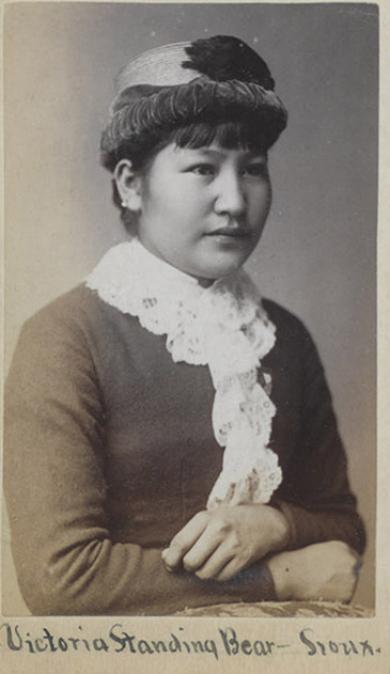 Black-and-white portrait of a young woman in Victorian-era clothing, adorned with beadwork and a lace scarf, labeled “Victoria Standing Bear-Conroy—Sioux: Beaded Legacies” at the bottom.