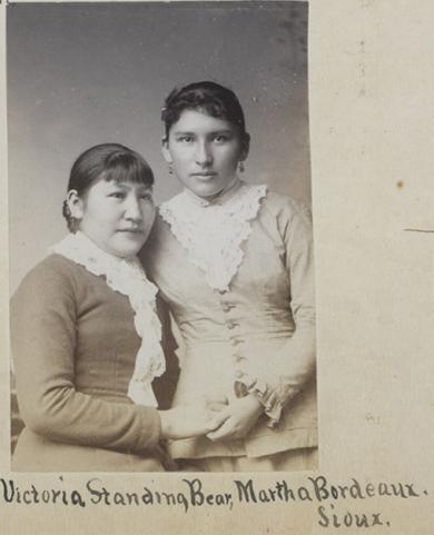 Two young women in 19th-century clothing pose for a studio portrait. Handwritten text identifies them as Victoria Standing Bear-Conroy and Martha Bordeaux, Sioux, whose elegant attire reflects the beaded legacies and artistry of their people.