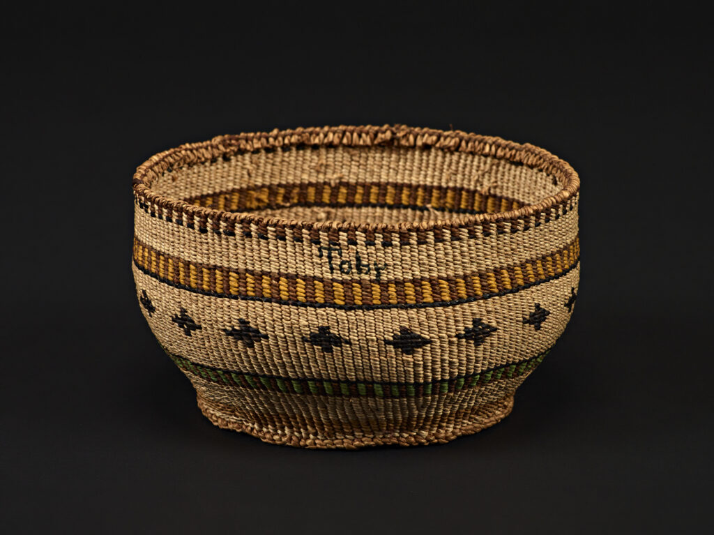A small basket with geometric patterns in brown, yellow, and green is displayed against a black background—one of our vault highlights that weaves together threads of big history and artistry.