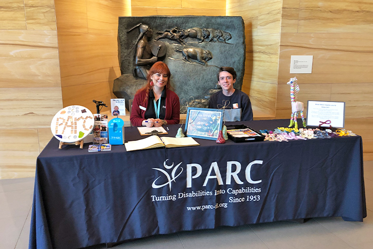 Two partners sit at a PARC informational table displaying brochures, crafts, and promotional items in front of a stone wall with a relief sculpture.