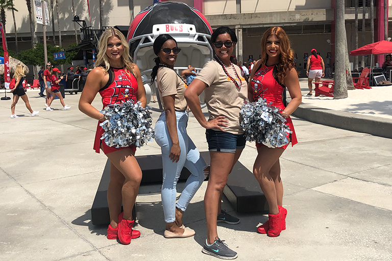 Two women pose with their partners, alongside two cheerleaders in red uniforms holding pom-poms, standing outside near a large football helmet display labeled "BULLS.