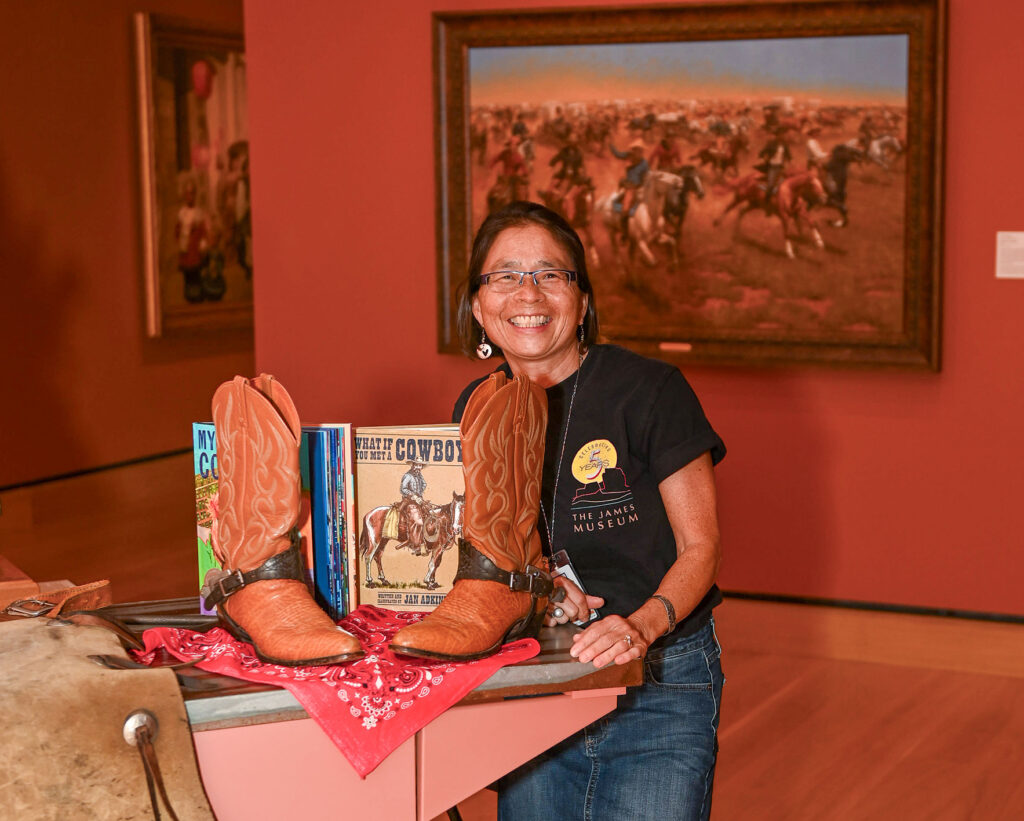 A woman stands next to a display featuring cowboy boots, books, and a red bandana in an art museum gallery with western-themed paintings on the wall.