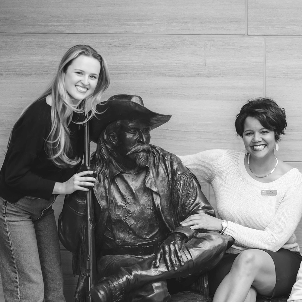 Two women pose and smile beside a seated bronze statue of a man holding a rifle, set against plain wooden paneling—an inviting example of unique decor often found in distinctive event spaces.