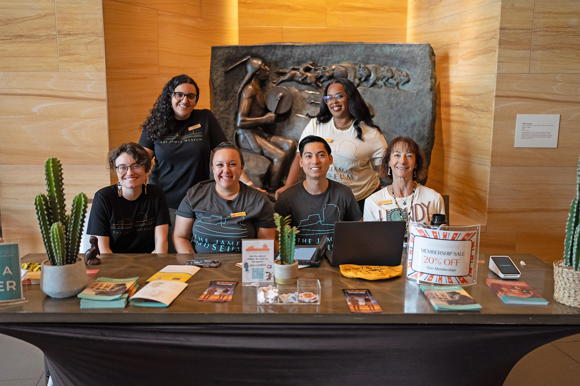 Six people sit and stand behind a museum information desk with brochures, a sign about a membership sale, and decorative cacti on display.