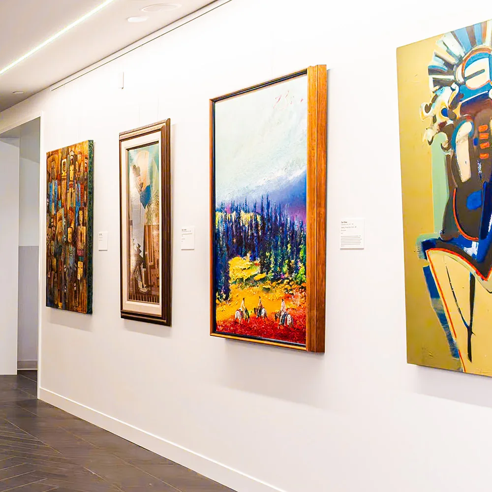 A row of framed paintings in various styles displayed on a white gallery wall with dark tile flooring, evoking the curated elegance of a Western Art Museum.