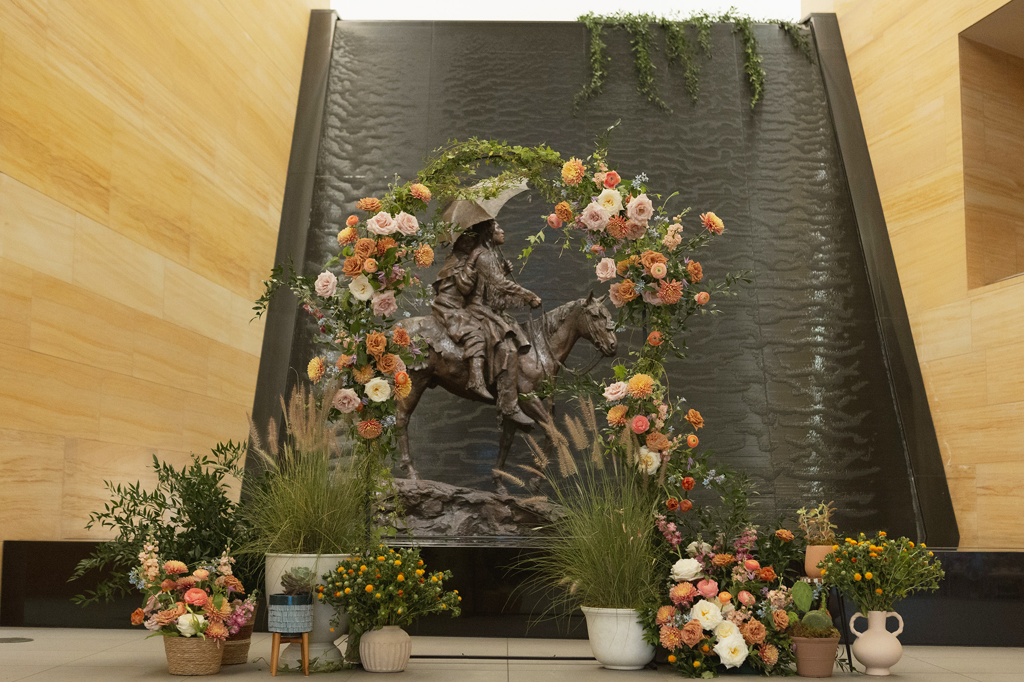A bronze statue of a woman on horseback is displayed indoors in front of a black water feature, surrounded by flowers and potted plants—an elegant centerpiece for sophisticated event spaces.